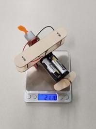 A wooden model airplane with batteries sits on a small digital scale. The scale reads 21.18 grams.