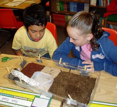 Photo of a boy and a girl looking at the sand, soil, bark, and water pool in the base of their model biodome.