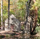 Photo shows a large wooden water wheel near a stone structure and creek.