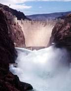 Photo looking upstream at the dam, a narrow point in a steep canyon filled with a concrete structure, holding back the river. Nearby, water pours from two outflow openings below the dam.
