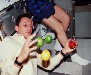 A photograph of an astronaut attempting to hold onto apples while in space. The apples are floating around the astronaut.