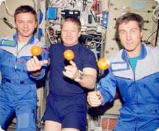 A photograph of three astronauts resting their right pointer fingers under oranges in a microgravity environment. The oranges are balancing on their fingers.