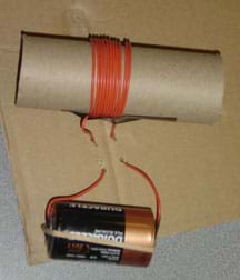 Photo shows a cardboard tube wrapped in wire and placed on a piece of flat cardboard. Two long tails of the coiled wire are woven through holes in the cardboard and are attached with a rubber band to opposite ends of a D-cell battery.