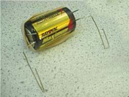 Photo shows the battery with a looped paperclip on each end. A rubber band stretches lengthwise across the battery to secure the two paperclips.