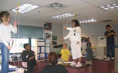 A photograph of many students standing on their desks in a classroom. The girls and boys are attaching their pulleys to the ceiling.