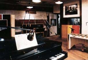 A photograph of a modern recording studio. Shown are a piano and microphones  in the foreground along with other musical instruments. 