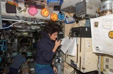 A picture of a female astronaut talking on a handheld device while floating in what looks like a scientific laboratory. Another astronaut in the background is collecting data on a computer.