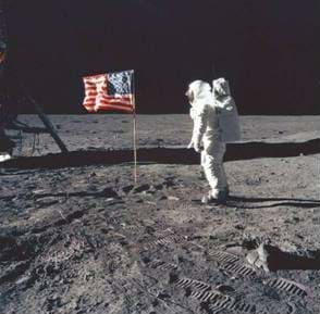 A photograph of Buzz Aldrin standing on the moon. He is standing next to the American flag.