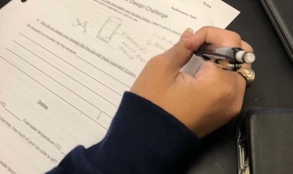 Close-up photo of a student’s right hand holding a pencil and writing on a piece of paper. The paper includes a drawing of a cup with labels around it. 