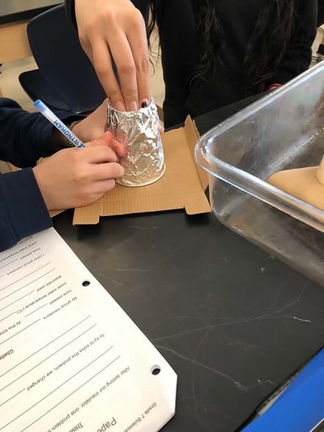 One student holds a foil-covered cup upside down on the table, while another student pushes the foil into the bottom of the cup.