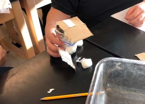 A student holds an insulated cup design. The paper cup is covered with some foil and cotton balls, and a cardboard lid has been added. 