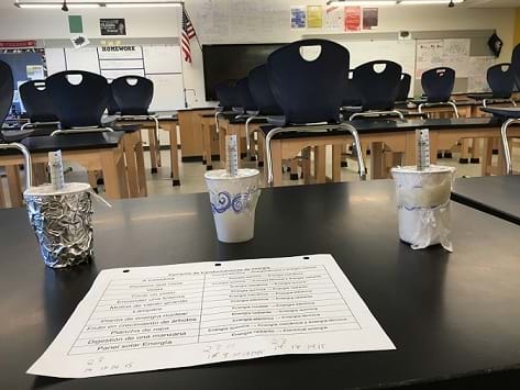 Three insulated cup designs with thermometers in them sit in a row on the table with a piece of paper in front of them. The left cup is covered in foil with a lid made of plastic wrap, and the center and right cups have lids made of plastic wrap.