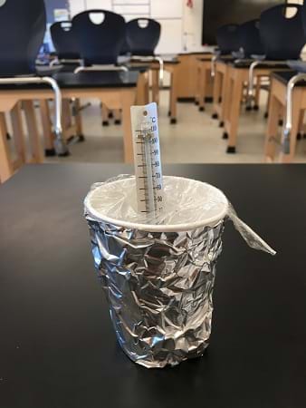 An insulated cup design filled with water sits on the table. The cup is covered in foil and has a lid of thin plastic wrap. A thermometer breaches the plastic lid and sits in the cup. 