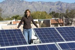 Photo shows a woman standing amidst dark blue angled solar panels on a city rooftop with a blue sky and mountain backdrop.