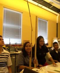 In a classroom, four students measure the height of a paper structure stretching from a table top to the ceiling.