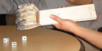 Photo shows a board for a forearm, and eyelet screws, fishing line, plastic tubing and duct tape combined to make a crude hand with fingers, along with dice at a table.