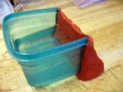 Photo shows a wedge of red clay that has replaced one side of a small plastic container, with the container filled with water to show that it holds water.