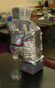 Photo shows a contraption made from a large tin can, plastic bottles and containers, aluminum foil and duct tape, sitting on a hot plate.