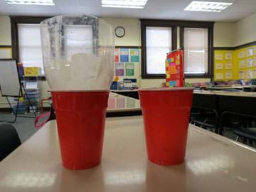 Photo shows two red cups, one with an upside-down top half of a two liter bottle containing a coffee filter resting inside of it.
