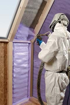 A man in white coveralls, gloves and a face mask sprays purple WALLTITE polyurethane spray foam insulation between the wooden stud framing of the walls of a house.