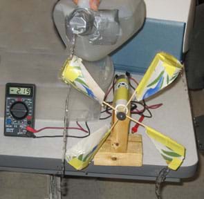Photo shows a hand holding a plastic gallon jug of water with the spout taped to restrict water flow and pouring water over paper cup turbine blades. A nearby multimeter reads 2.19.