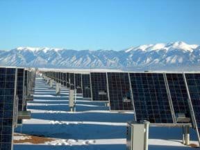 Photo shows endless rows of angled photovoltaic panels mounted on a snow-covered field with a background of snowcapped mountains and blue skies.