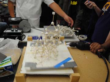 On a tabletop surrounded by students, two framework structures made of toothpicks and mini marshmallows jiggle on moving platforms; one structure has collapsed.