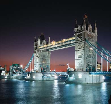 Dusk photo shows lighted two-tower bridge with cables.