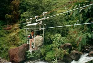 Photo shows two people sitting and facing each other in a metal-cage cable car that is hanging from pulleys resting on a steel wire strung across a river.