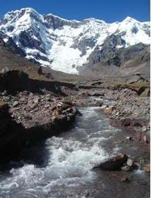 Photograph of a large mountain covered in snow (glacier) with a glacial stream flowing down valley of the mountain. 