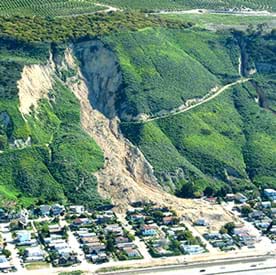 A photograph shows a large landslide caused by water erosion. A town at the base of a mountain is partially destroyed. Many of the town's structures are completely or partially covered by dirt from the mountain landslide that came down from above it.