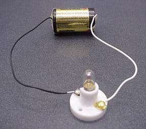 Photo shows a D-cell battery with a white wire connecting the positive terminal of the battery to one terminal of a light bulb holder, and a black wire connecting the negative terminal of the battery to the opposite terminal of the same light bulb holder.