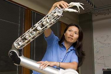 A photograph of a woman standing in front of a prosthetic arm controlled using robotics.