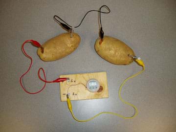 A photograph shows two potatoes with pennies and nails halfway embedded. Alligator clips are attached to each penny and nail, and connected in series to power a small digital clock.