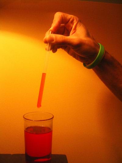 A photograph shows finger plugging the top of a clear straw, causing red-colored water to stay in the straw when it is lifted out of a glass.