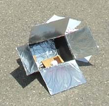 A photo shows a box with flaps covered in foil, and chocolate and marshmallows on a graham cracker inside the box.