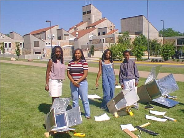 A photo shows four teens and three shiny, box-type solar ovens in a sunny field on a university campus.  