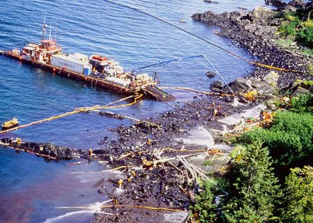 A photograph shows beach workers and a maxi-barge joining forces to clean a gunky black beach after the Exxon Valdez oil spill.