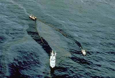 An arial photograph shows two boats dragging a net through the ocean waters, skimming oil off the water surface.