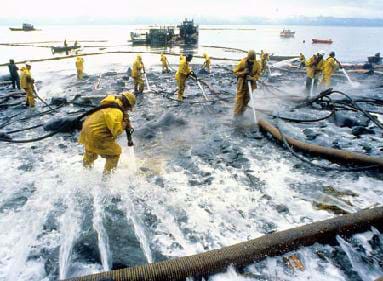 A photograph shows people in yellow protective clothing along the sandy shoreline hauling hoses and spraying oiled rocks with high pressure water.