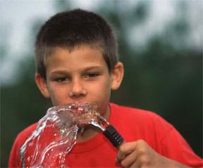 A photograph of a small boy, wearing a red t-shirt, drinking water from a garden hose.
