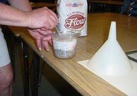 A photograph showing a glass measuring cup full of pebbles, water and flour. A person is stirring the mixture. Also shown are a large white funnel and a bag of flour.