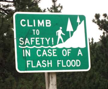 A photograph shows a green and white road sign with the message: Climb to safety in case of a flash flood.