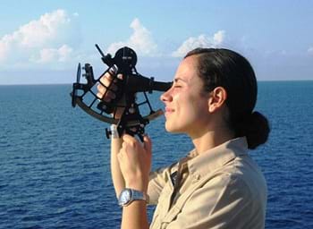Photo shows a woman on a boat in the ocean using a sextant.
