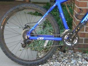 A photograph of a bicycle, showing the rear half of the frame, its wheel, tire, gears and spokes.