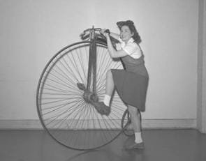 A black and white photograph of a girl getting on an old bicycle. The bicycle has one big front wheel and a small back wheel.