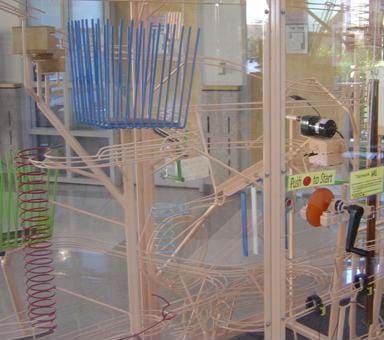 Photo shows a Rube Goldberg machine, a complex system of loops and tracks to simply move a ball throughout a large plastic-enclosed box.