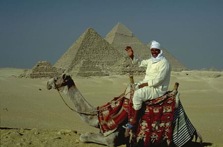 Photograph of a man on a camel in front of five pyramids in a desert.