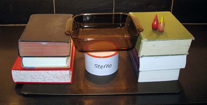 A glass baking dish is suspended by stacks of three books on each side over a can of sterno. Food coloring bottles are nearby.