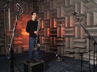 Photo shows a man playing a saxophone surrounded by microphones in a room with walls covered in acoustical tiles.